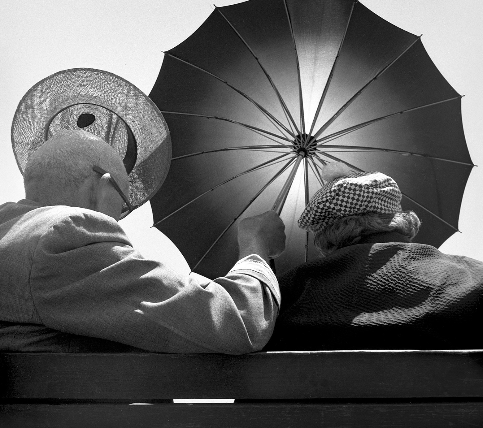Two old people seen from behind sitting on a bench. The man on the left is wearing a coat and taking his hat off his head with his left hand as he holds an umbrella in his right hand blocking the sun. The person on the right is wearing a hat and a coat.